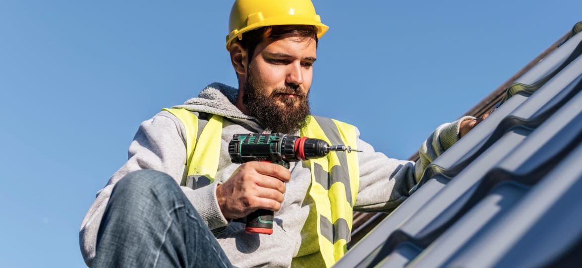 Construction worker in safety gear installing roofing materials with a power drill, against a clear blue sky, emphasizing roof renovation services.