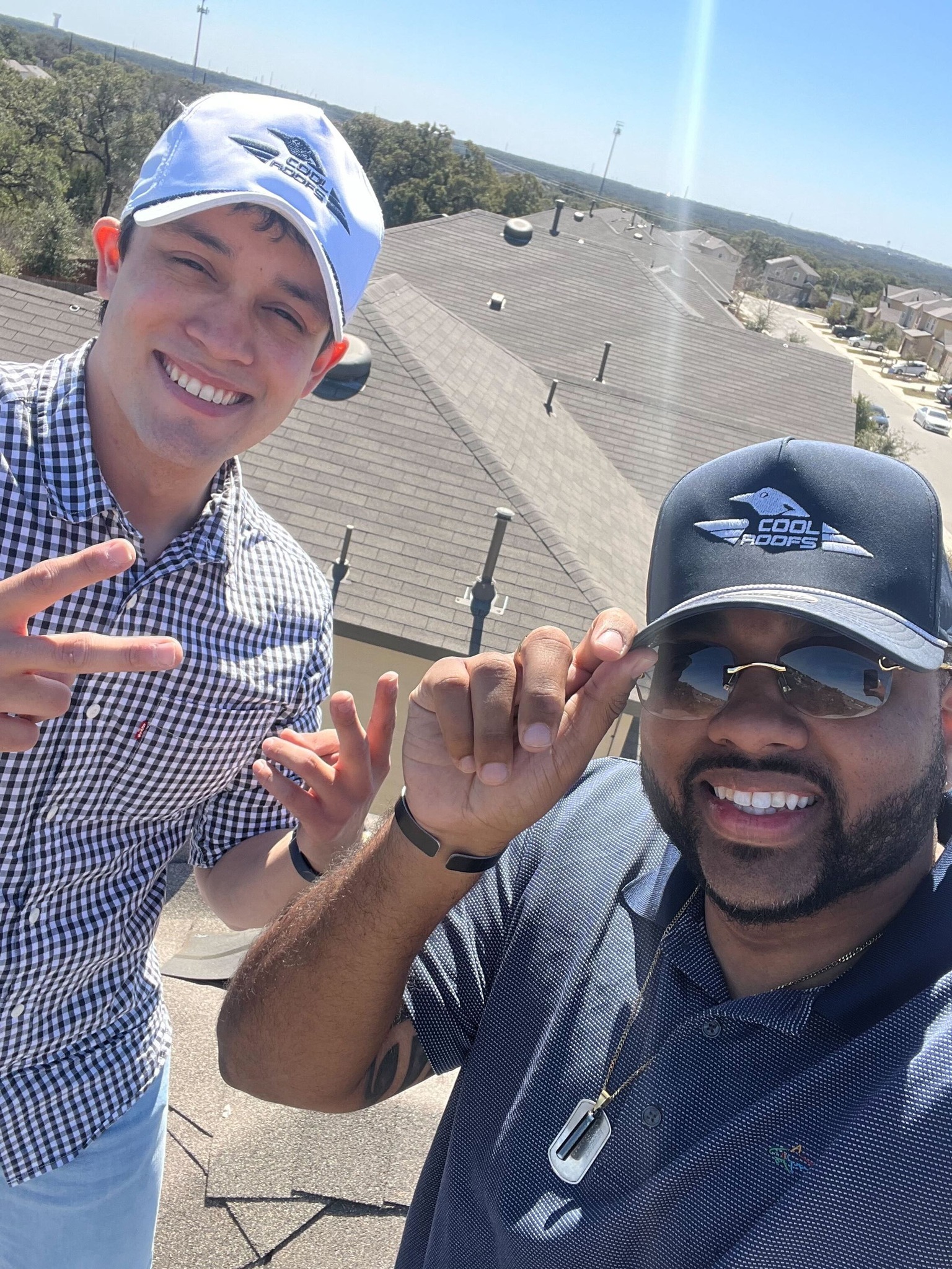 Two smiling men on a rooftop wearing Cool Roofs hats, showcasing a friendly atmosphere while conducting a roof inspection.