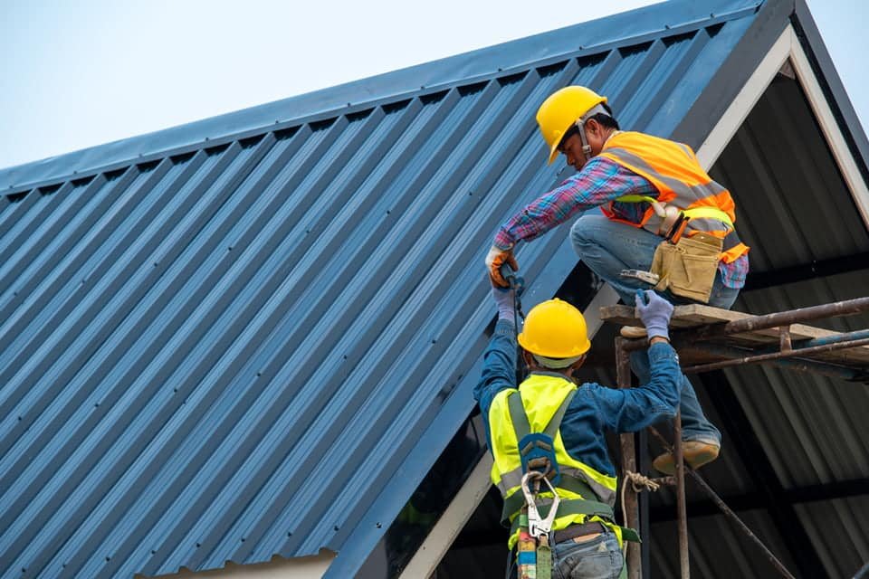 Two construction workers installing a commercial metal roof, wearing safety helmets and vests, on a building, emphasizing quality workmanship and compliance with industry standards.
