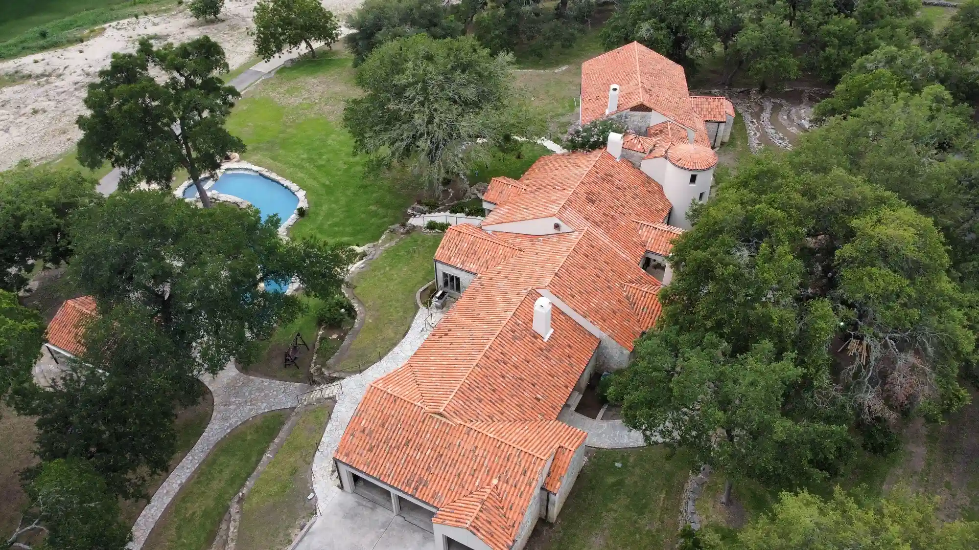 Aerial view of a residential property with a red-tiled roof, surrounded by green trees and a swimming pool, illustrating a potential site for roof leak repair services in Texas.