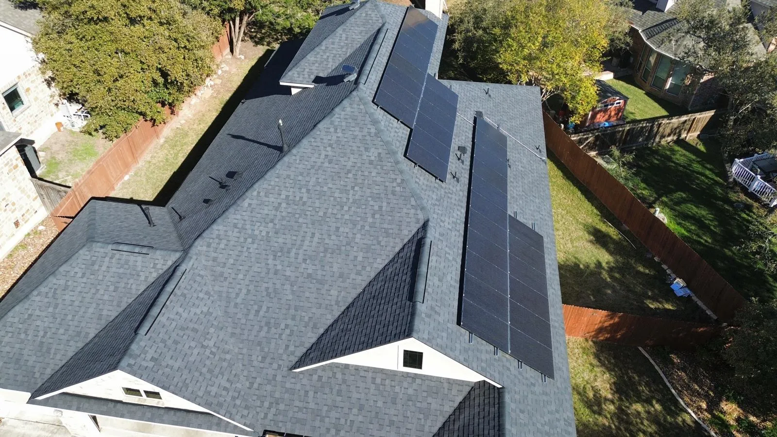 Roof with dark shingles and solar panels installed, surrounded by trees and a fenced yard, illustrating energy-efficient roofing solutions relevant to Texas homeowners.